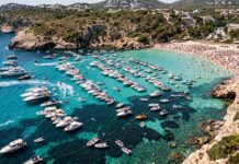 Summer migrations are breaking the Mediterranean: How overtourism destroys marine climate defenses Una baia del Mediterraneo completamente invasa da imbarcazioni ancorate vicino alla costa.