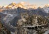 Leopardo delle nevi accovacciato su una scogliera rocciosa delle montagne dell'Himalaya con vette innevate sullo sfondo